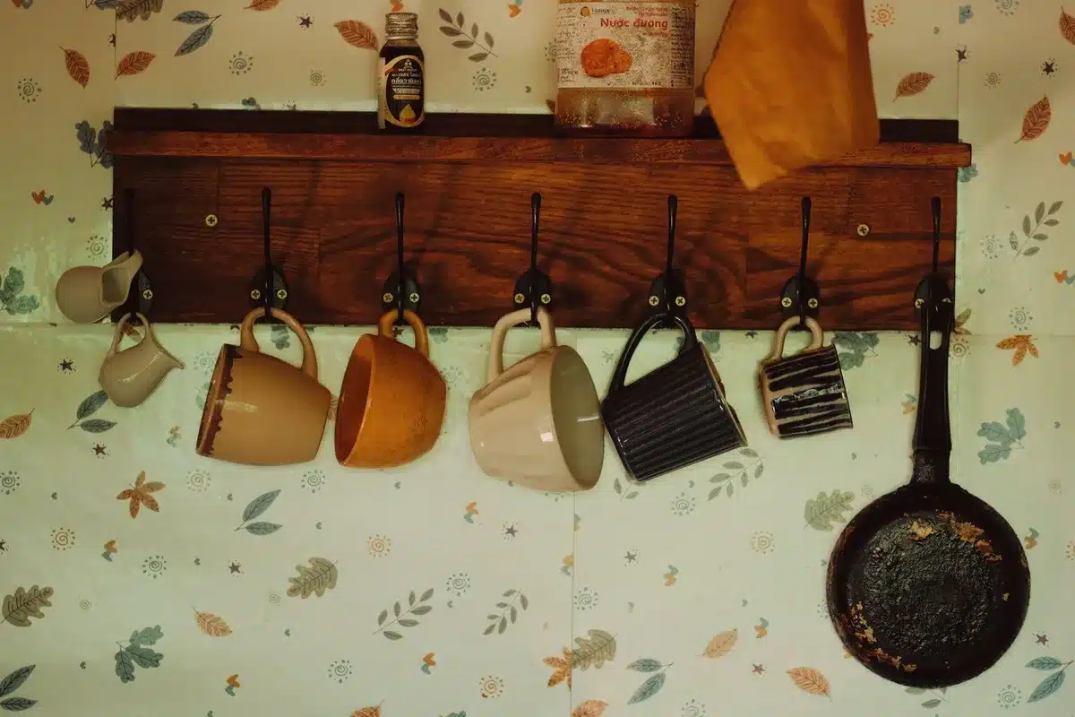 Cordless drill, measuring tape, wall anchors, and screw-in mug hooks arranged on a wooden work surface.