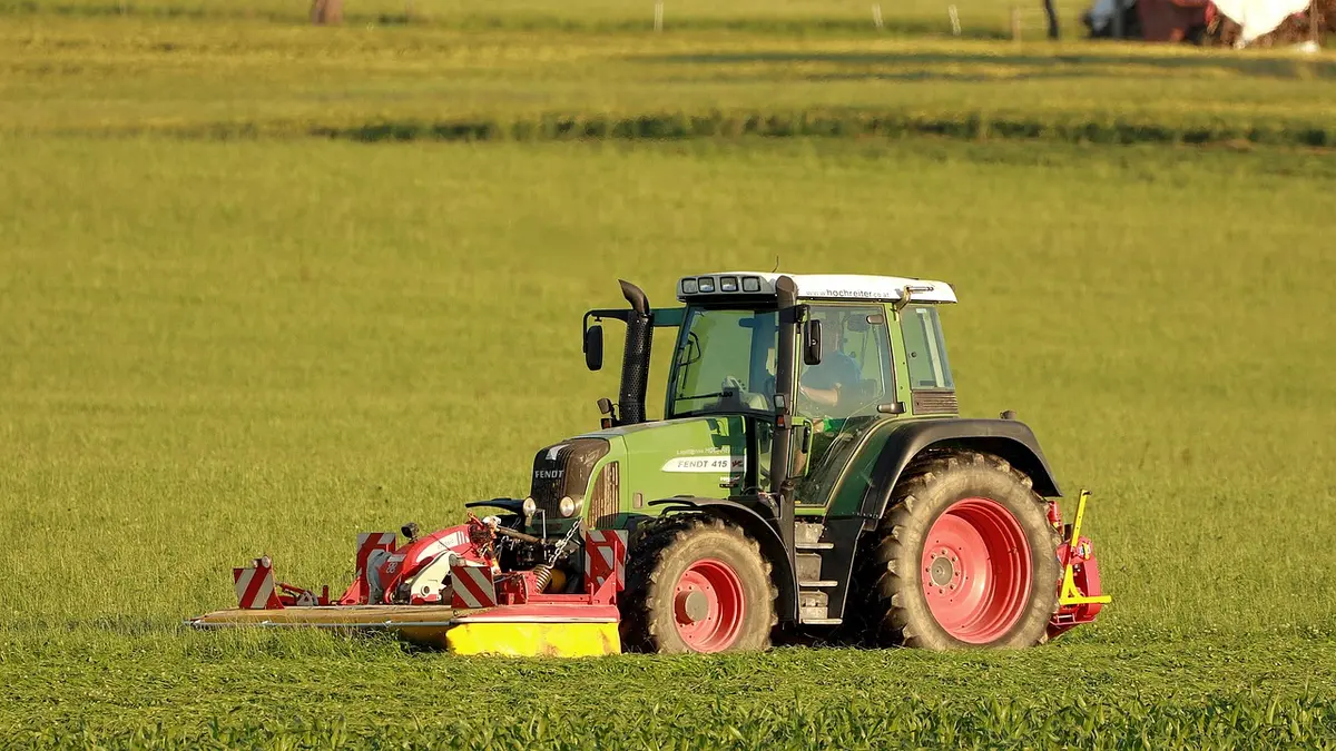 Gloved hands adjusting a mower wheel lever under the deck with clippings on lawn
